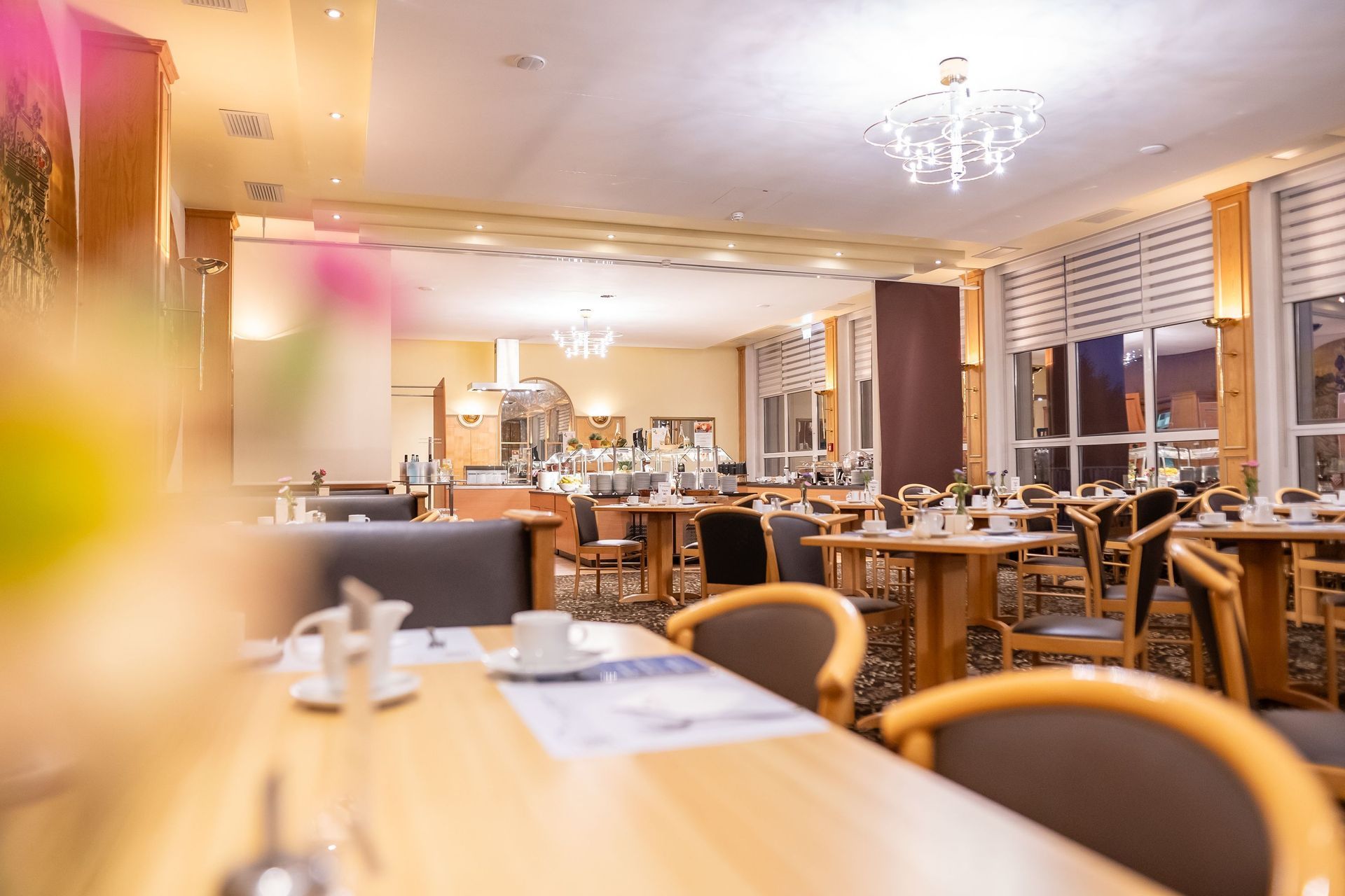 Warm beleuchteter Speisesaal eines Restaurants mit Tischen, Stühlen und Kronleuchtern, gesehen von einem leicht verschwommenen Tisch im Vordergrund.