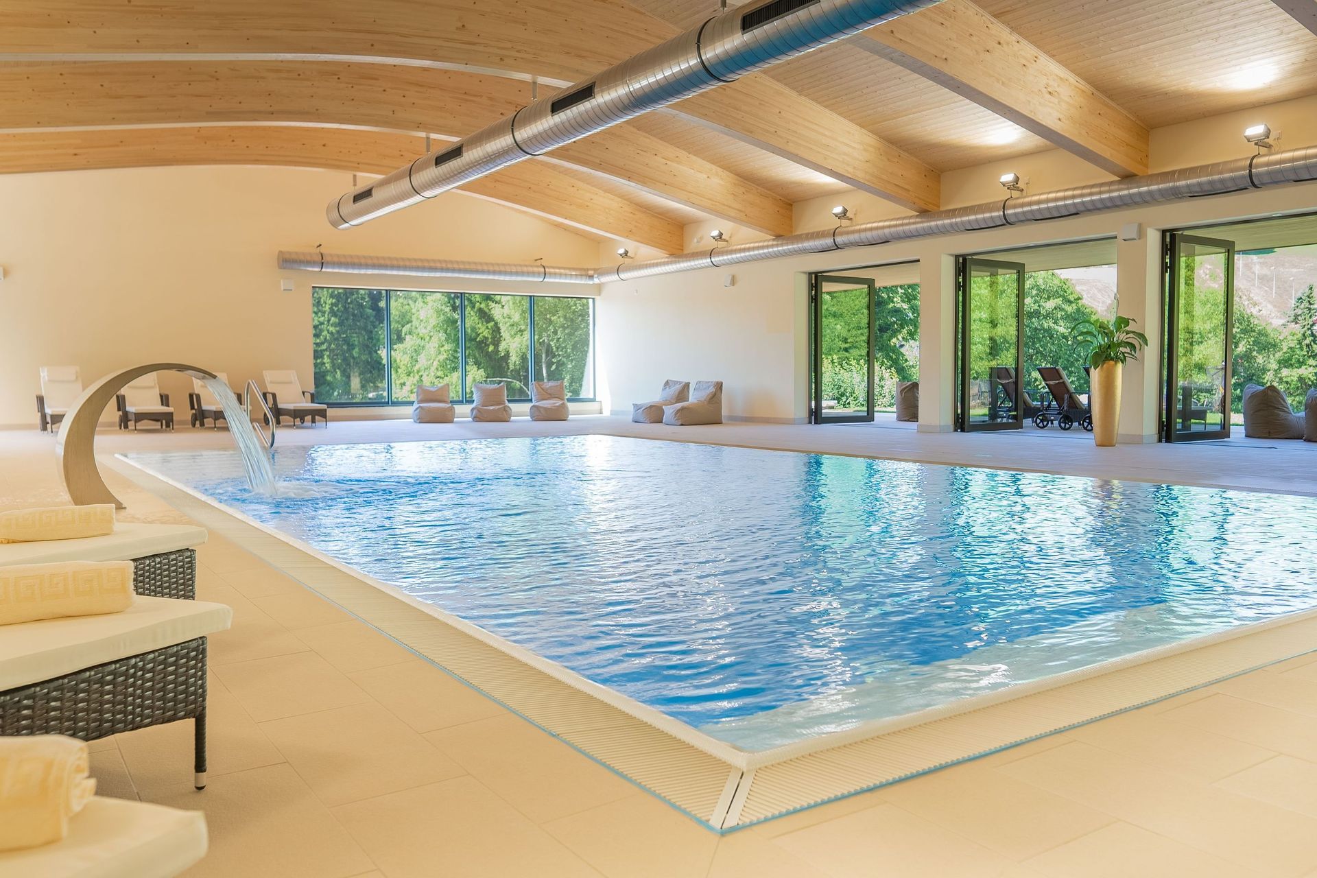 Hallenbad mit klarem, blauem Wasser, beiger Terrasse und großen Fenstern mit Blick ins Grüne.