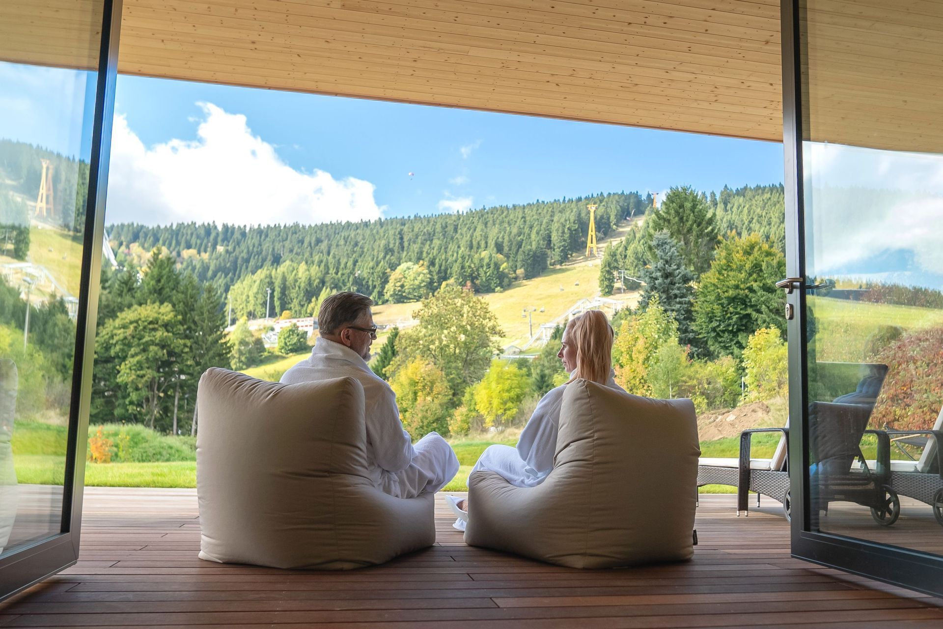 Zwei Personen sitzen auf Sitzsäcken und genießen durch ein großes, offenes Fenster den Blick auf die sonnige Berg- und Waldlandschaft.