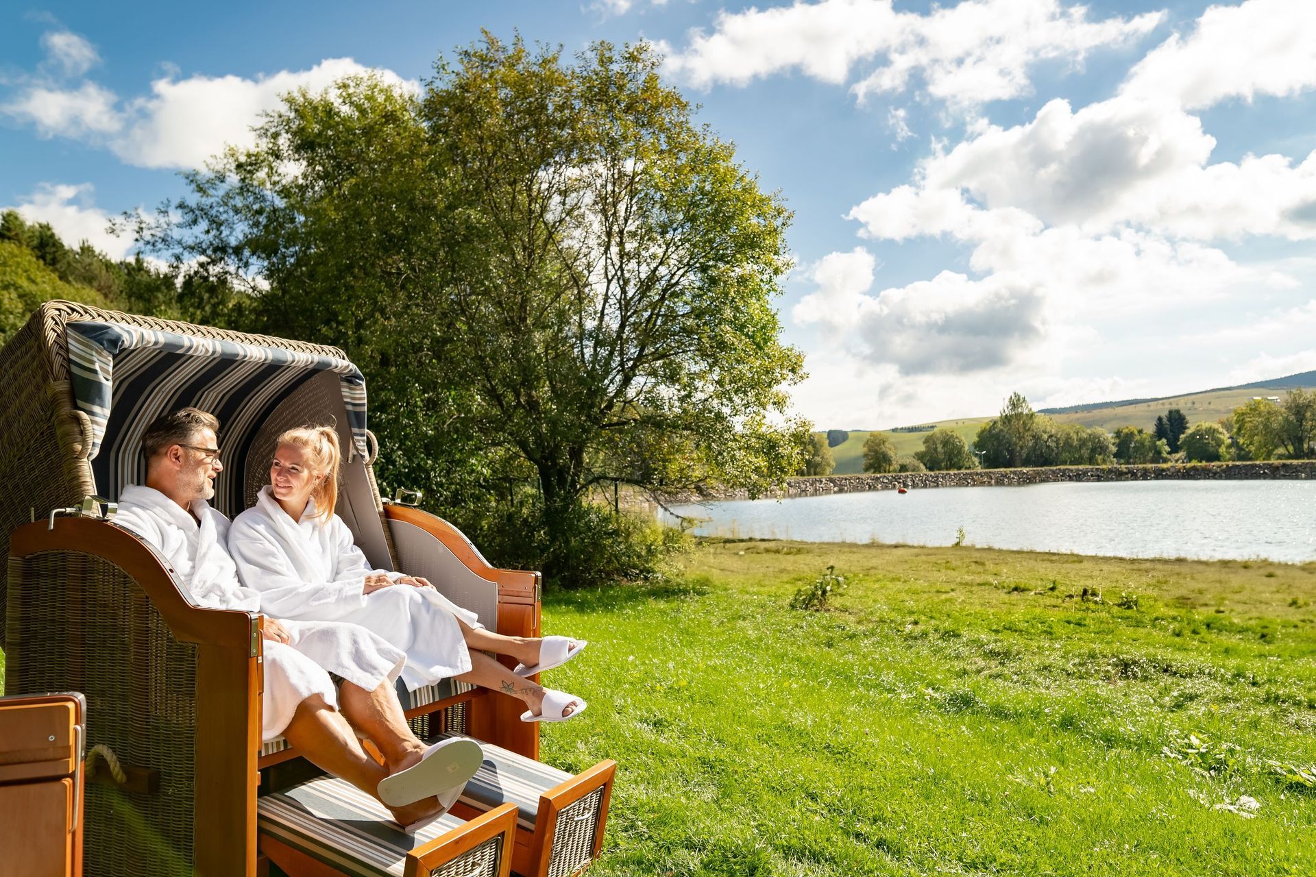 Zwei Personen entspannen sich in Korbstühlen an einem See auf einer sonnigen Rasenfläche.