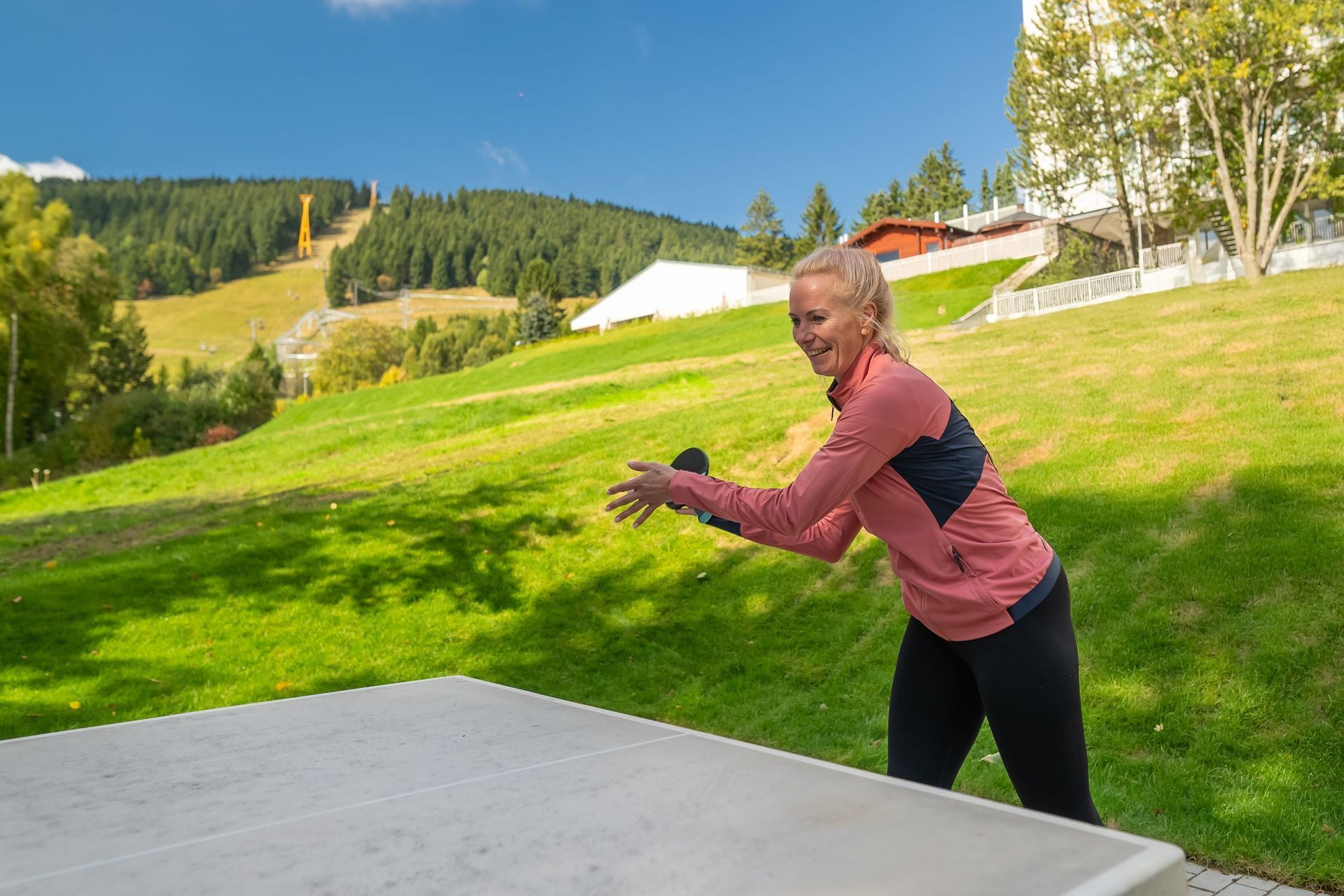 Eine Frau wirft eine Frisbee an einem grasbewachsenen Hang; im Hintergrund sind Berge und Bäume zu sehen.