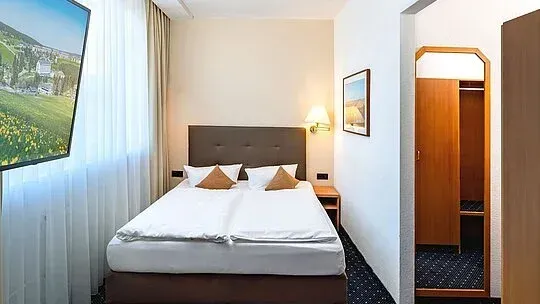 Kleines Hotelzimmer mit zwei Betten, einem an der Wand montierten Fernseher und einem Fenster mit dünnen Vorhängen.