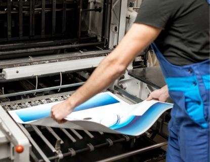 Person in blue overalls handling printed sheet from a printing press, showcasing blue and white design.