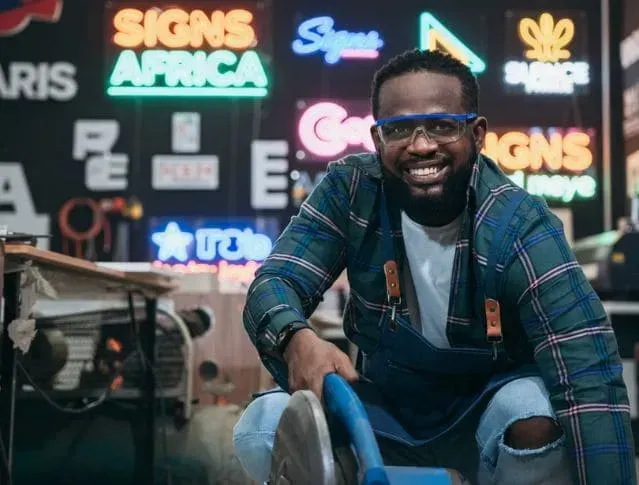 Man in workshop smiles, holding saw. Neon signs in background.