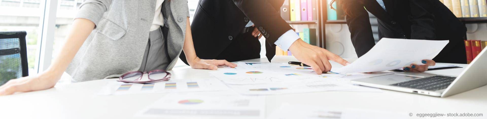 Viele ausgebreitete Dokumente auf dem Tisch. Eine Person zeigt auf etwas und eine andere Person steht daneben.