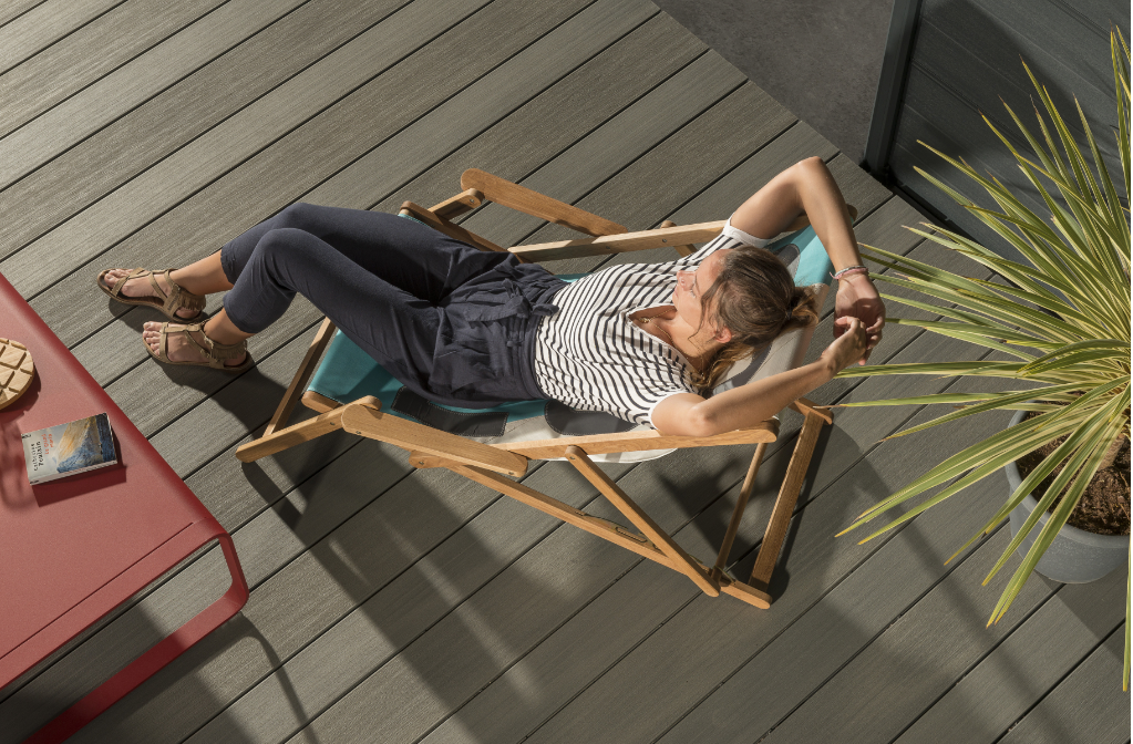 Une personne allongée sur une chaise en bois sur une terrasse grise, à côté d'une table rouge et d'une plante verte.