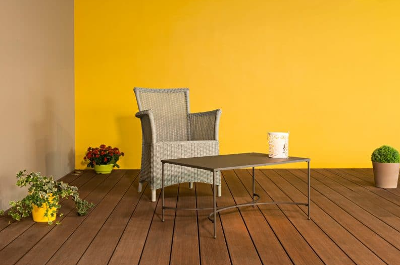 Chaise en osier et petite table sur une terrasse en bois, adossée à un mur jaune vif, avec des plantes en pot.