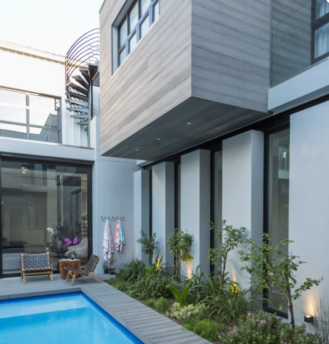 Patio de maison moderne avec piscine bleue, murs de verre et petits aménagements paysagers
