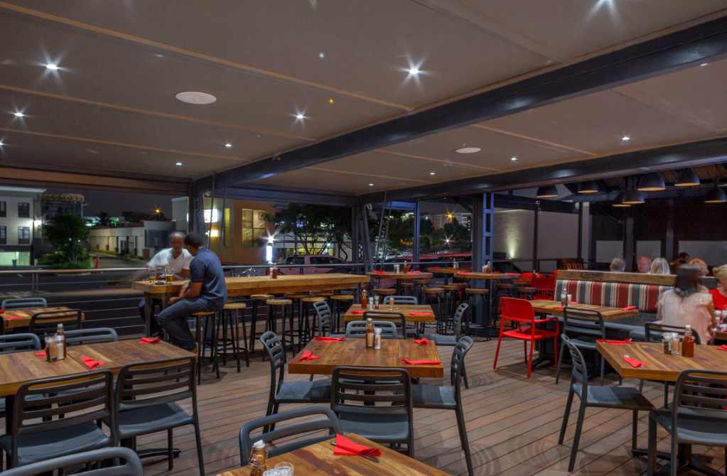Terrasse de restaurant en plein air avec tables et chaises vides sous un auvent illuminé, deux personnes assises au loin