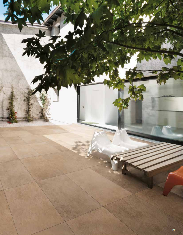 Patio moderne avec carrelage beige, chaise longue blanche, banc à claire-voie, paroi vitrée et arbre feuillu au-dessus.