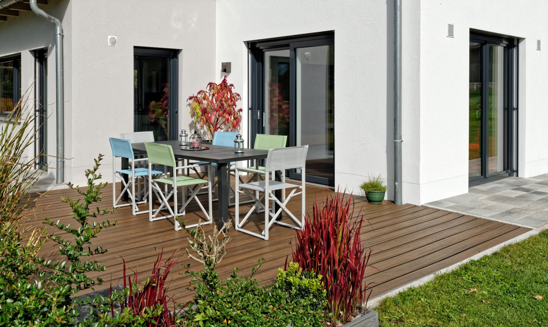 Patio moderne avec table à manger extérieure, chaises, plantes en pot et une maison blanche aux grandes portes vitrées