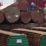 Des barils métalliques rouillés empilés et des tuyaux en fagots dans une zone industrielle.