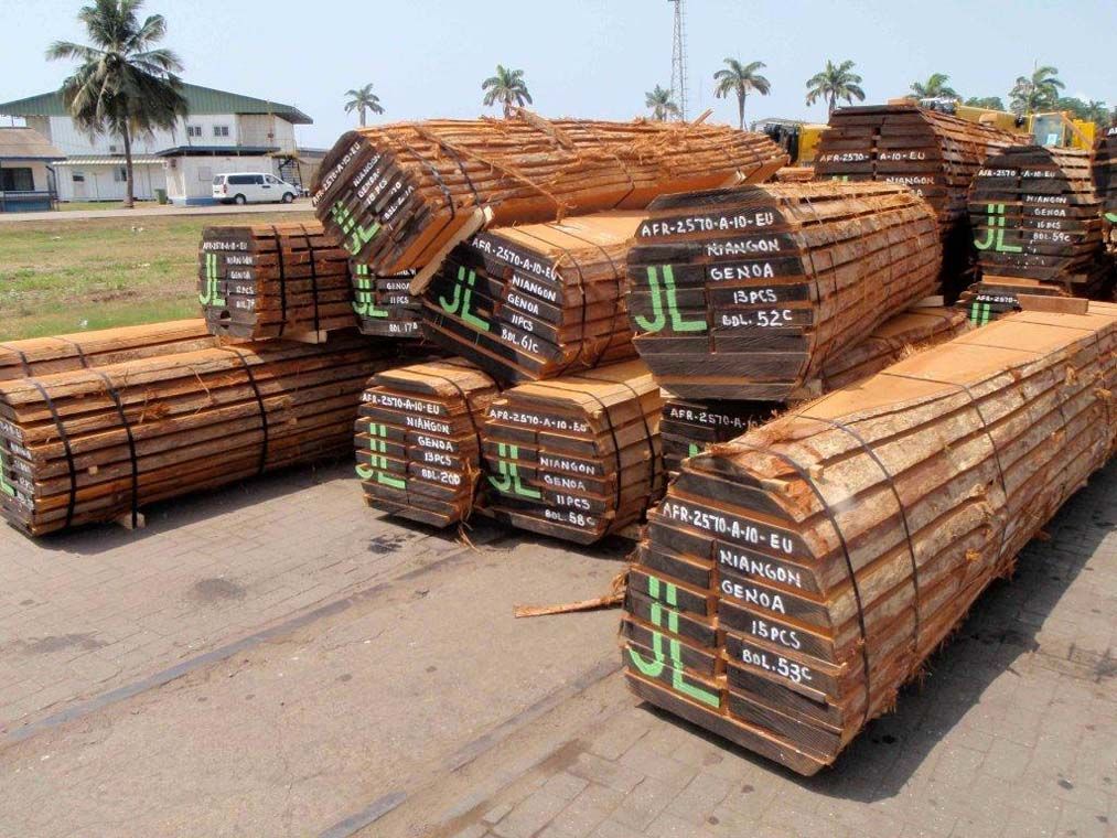 Des piles de tuyaux en bois ou de barres d'armature, entassés en bord de route, avec des palmiers et des bâtiments en arrière-plan.