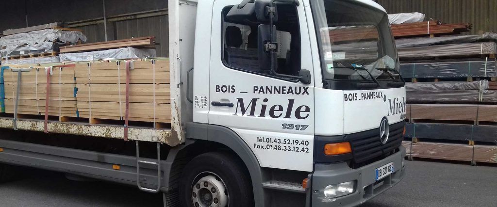 Camion plateau blanc chargé de caisses en bois, stationné à l'intérieur d'un entrepôt, sur un quai de chargement.