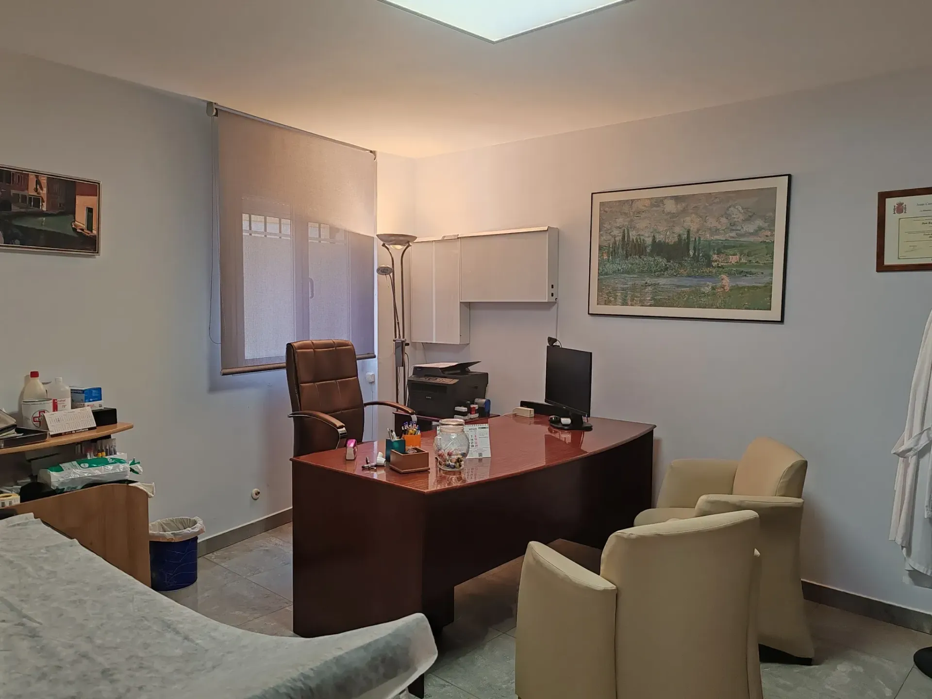 Interior del consultorio médico con escritorio, sillas, equipo médico y una ventana con persianas.