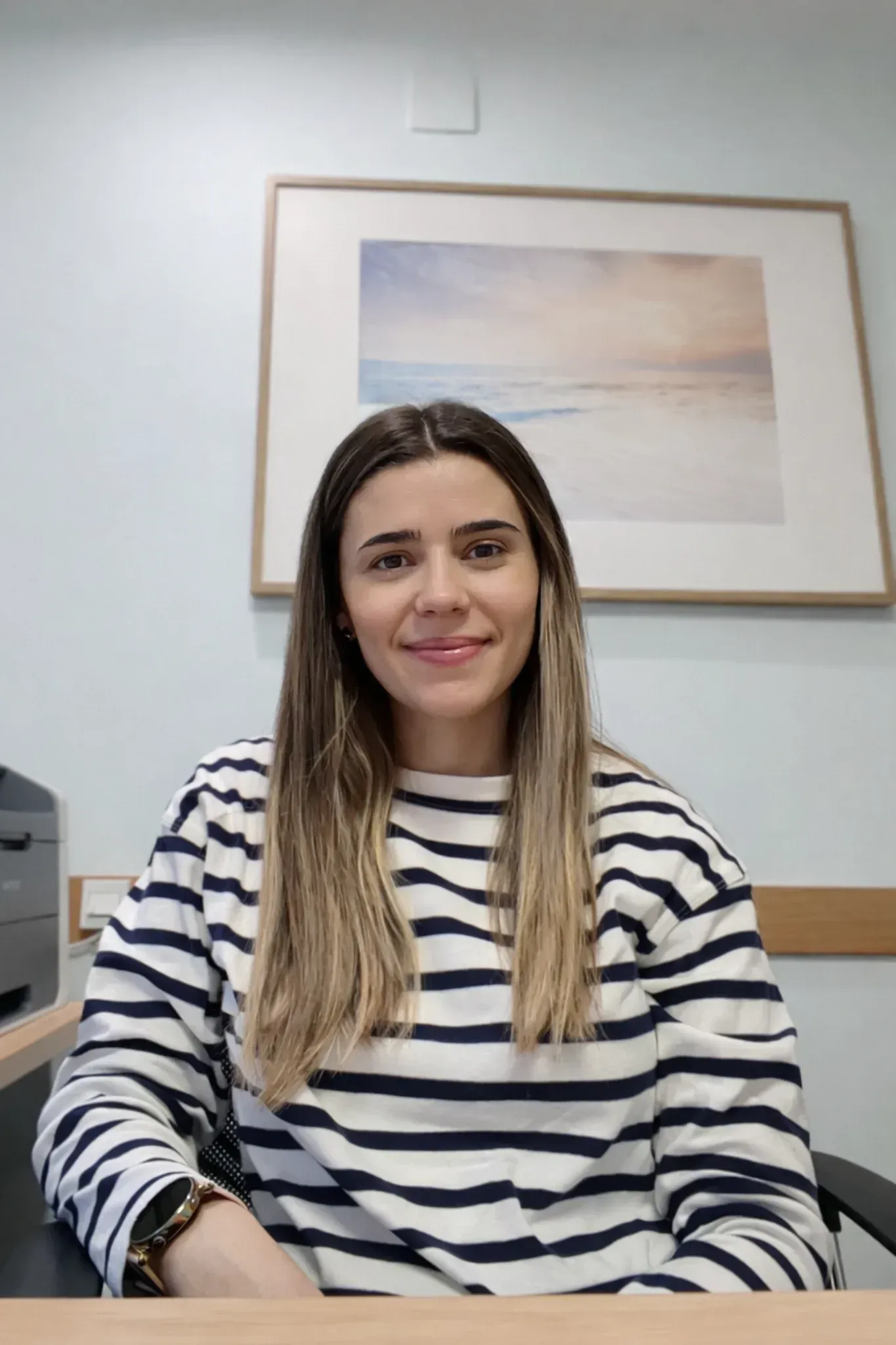 Mujer con camisa de rayas sonriendo, sentada en un escritorio con una imagen de una escena de playa en la pared.