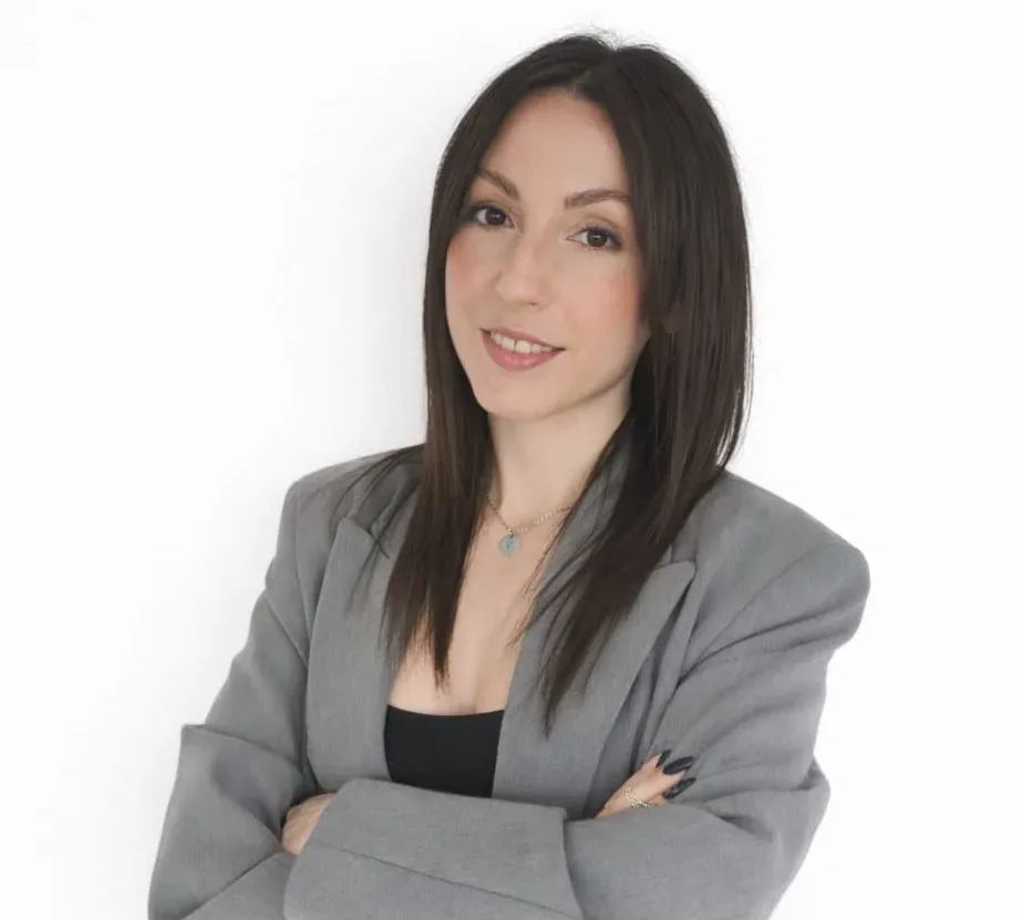 Mujer con cabello largo y oscuro, vestida con un blazer gris, con los brazos cruzados, sonriendo, sobre un fondo blanco.