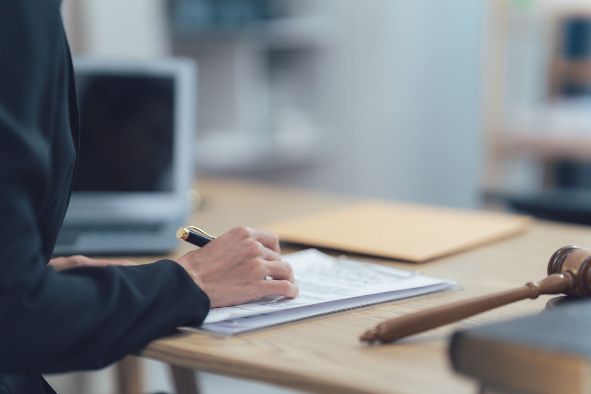 Une personne en costume rédigeant un document à un bureau avec un maillet, un livre et un ordinateur portable.