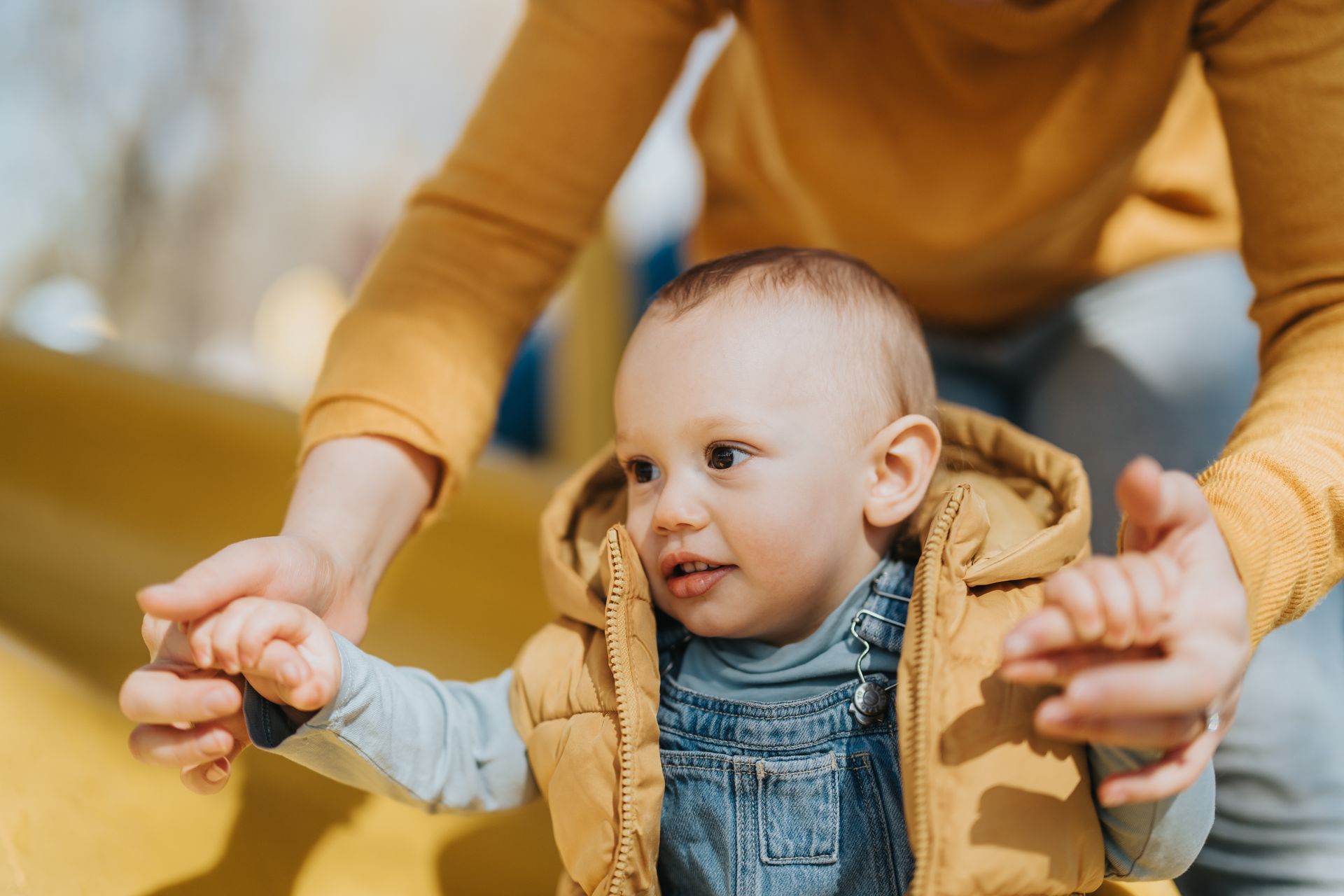 Enfant en gilet jaune et salopette, aidé par un adulte à descendre un toboggan jaune.