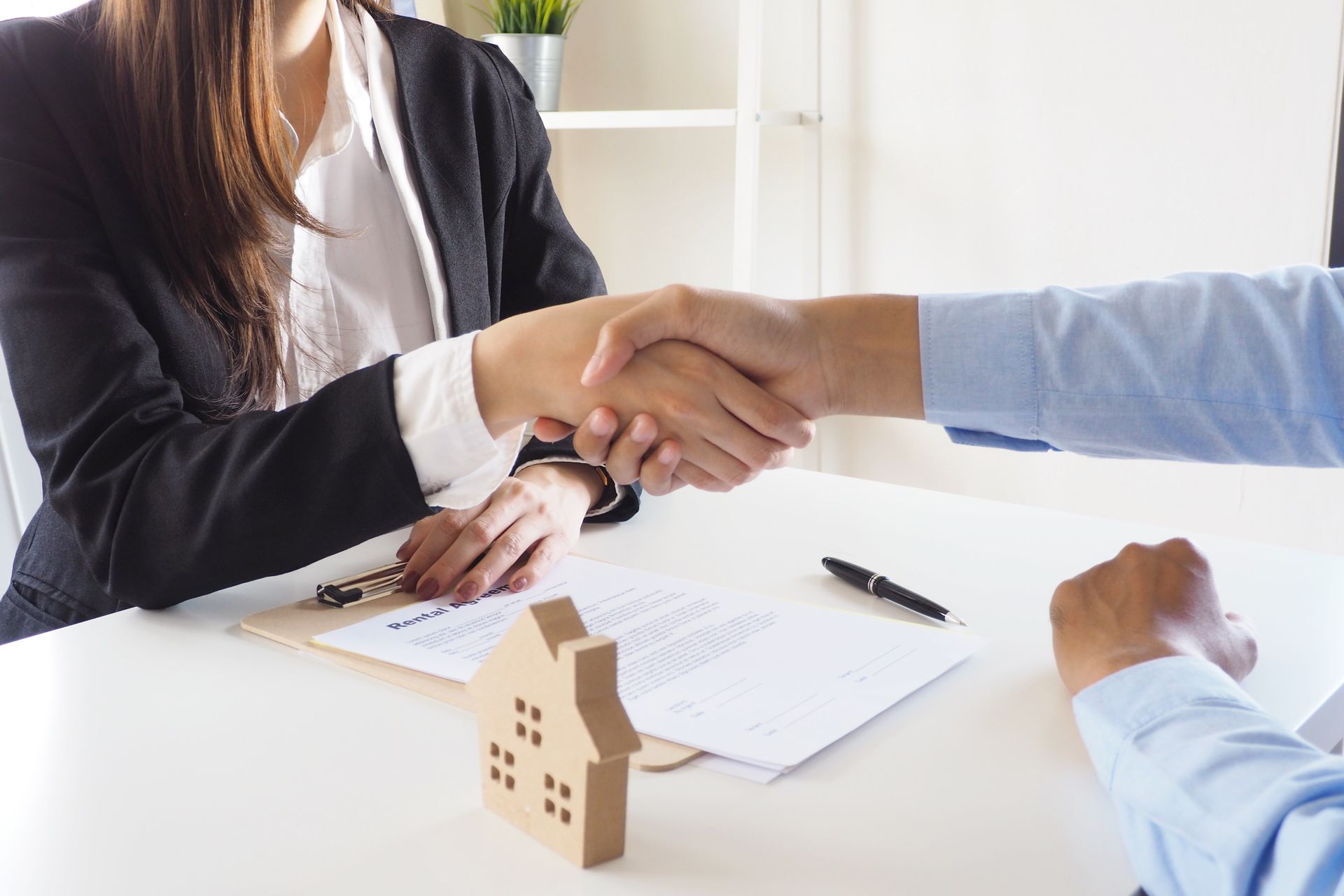Deux personnes se serrent la main au-dessus d'un contrat immobilier posé sur un bureau, une maquette de maison en bois à proximité.