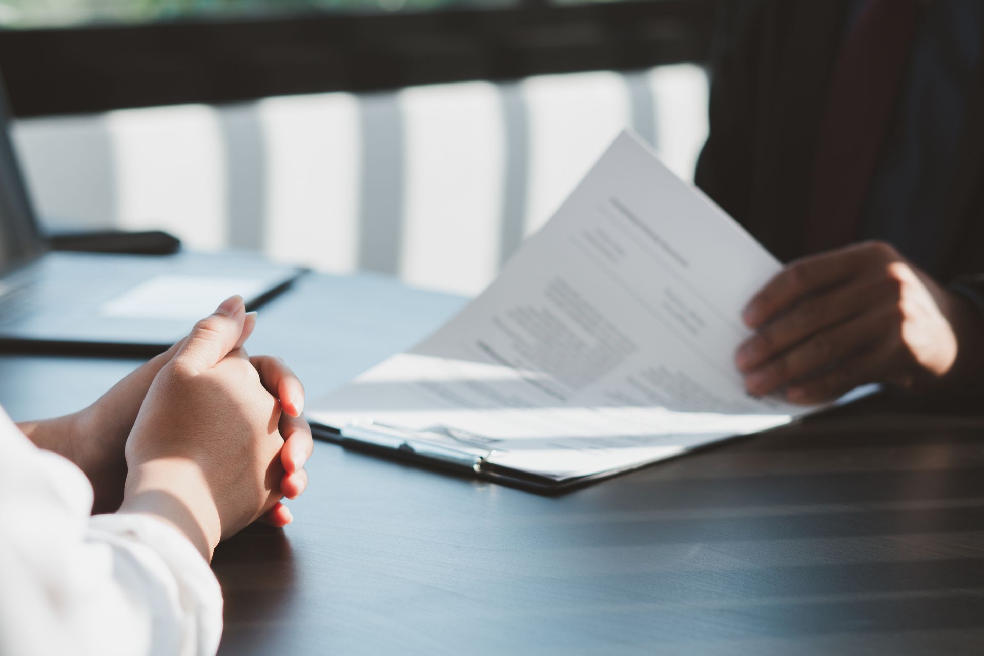 Les mains d'une personne sont jointes sur un bureau, face à une autre personne tenant des papiers.