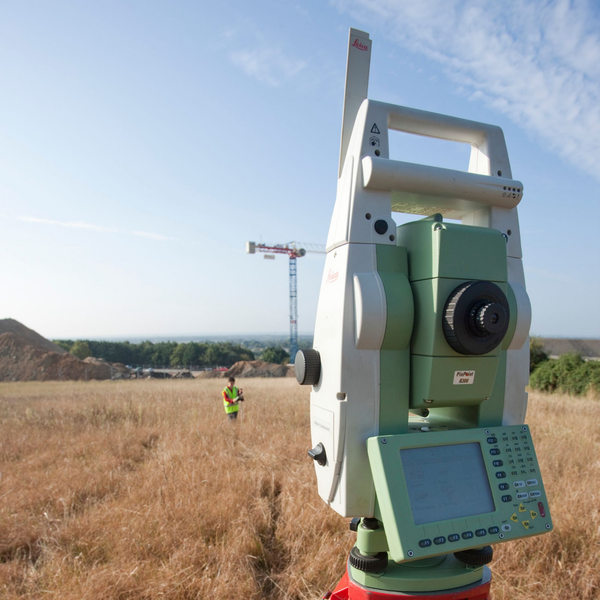 Du matériel topographique sur un trépied dans un champ, avec une personne au loin et une grue visible sous un ciel bleu.