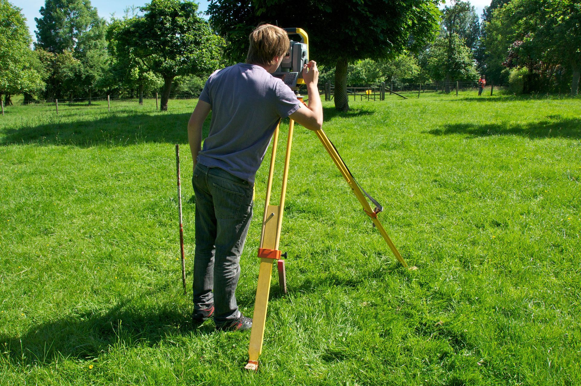 Un homme utilise un instrument d'arpentage sur un trépied, à l'extérieur, dans une zone herbeuse.