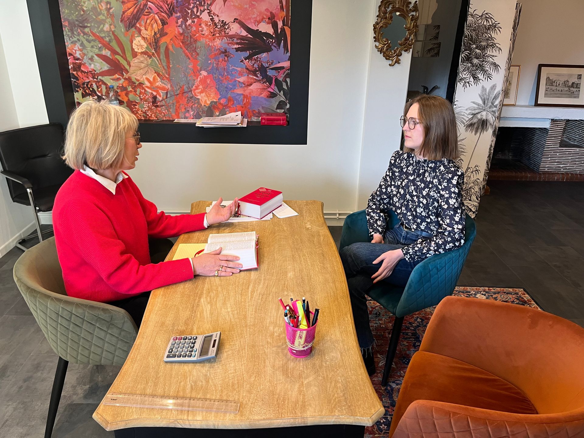 Deux femmes assises à une table, probablement en réunion ; l'une porte un pull rouge, l'autre une chemise à motifs.