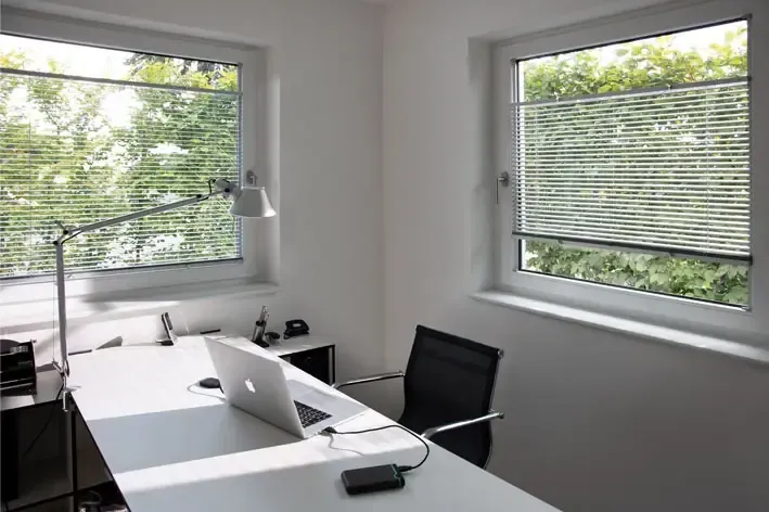 Ein Schreibtisch mit einem Laptop und einer Lampe darauf vor einem Fenster.