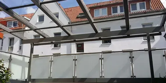 Ein Balkon mit Glasdach und Edelstahlgeländer vor einem Gebäude.