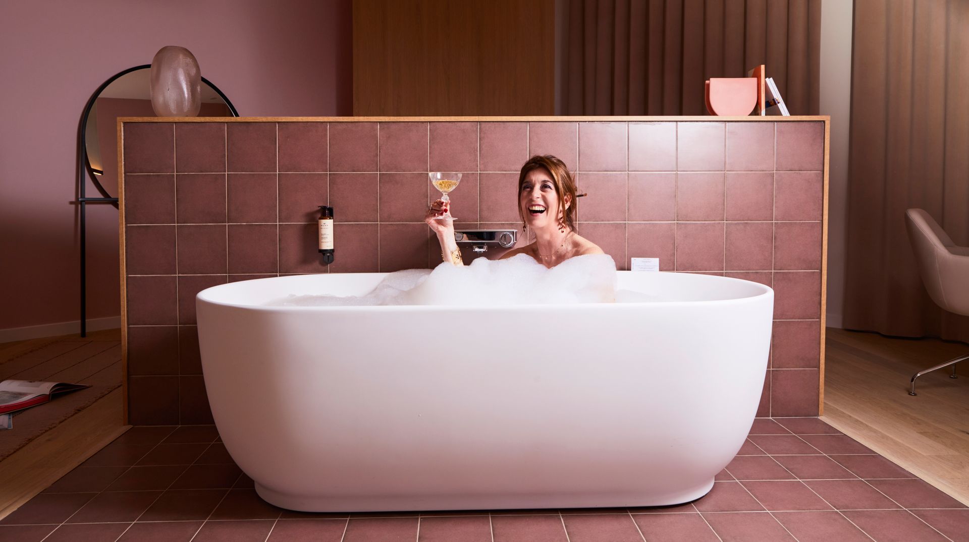 Une personne se détend dans une baignoire blanche, dans une salle de bains moderne carrelée de rose, un verre à la main.