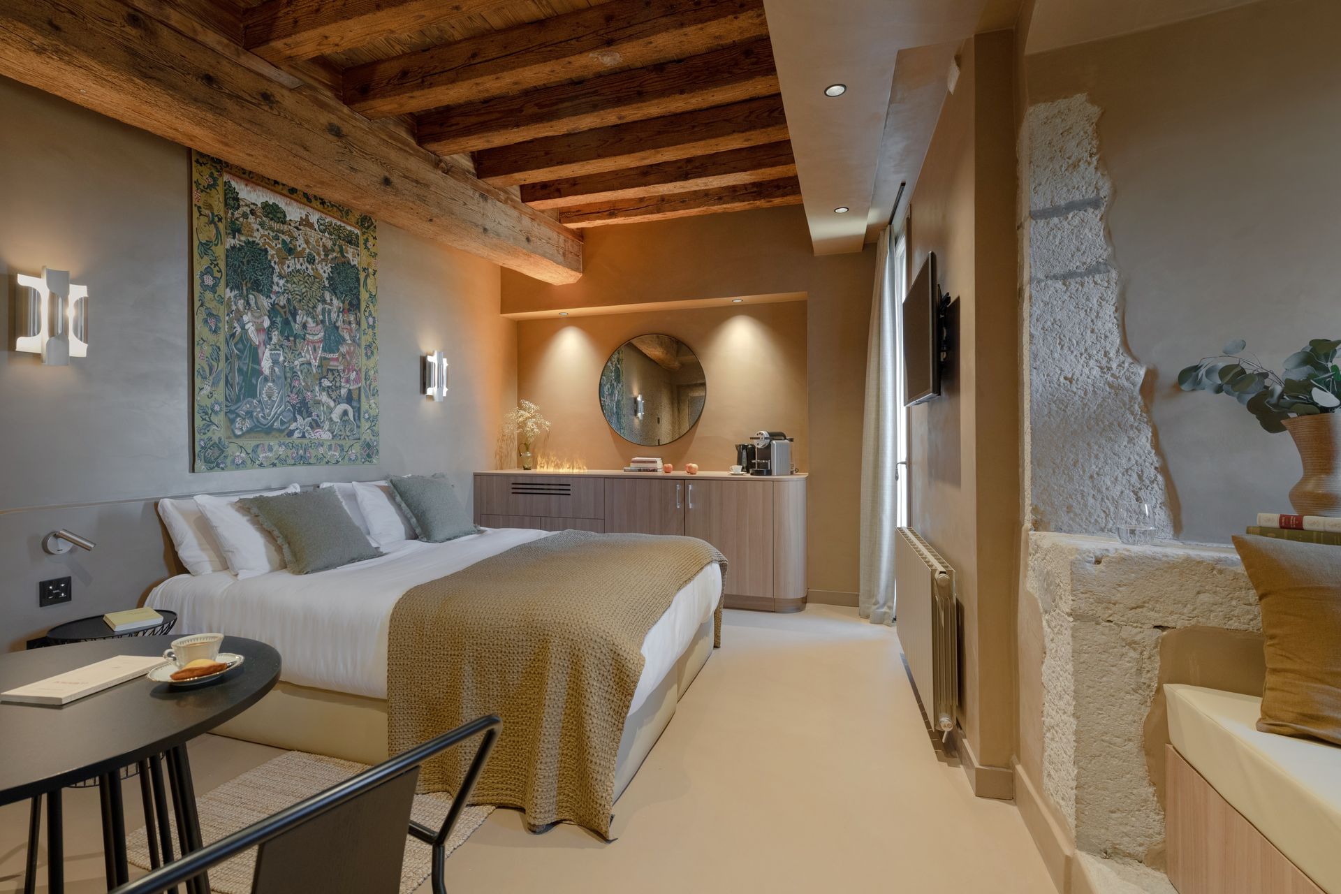 Chambre avec poutres apparentes, tapisserie, lit, miroir rond et mur en pierre.