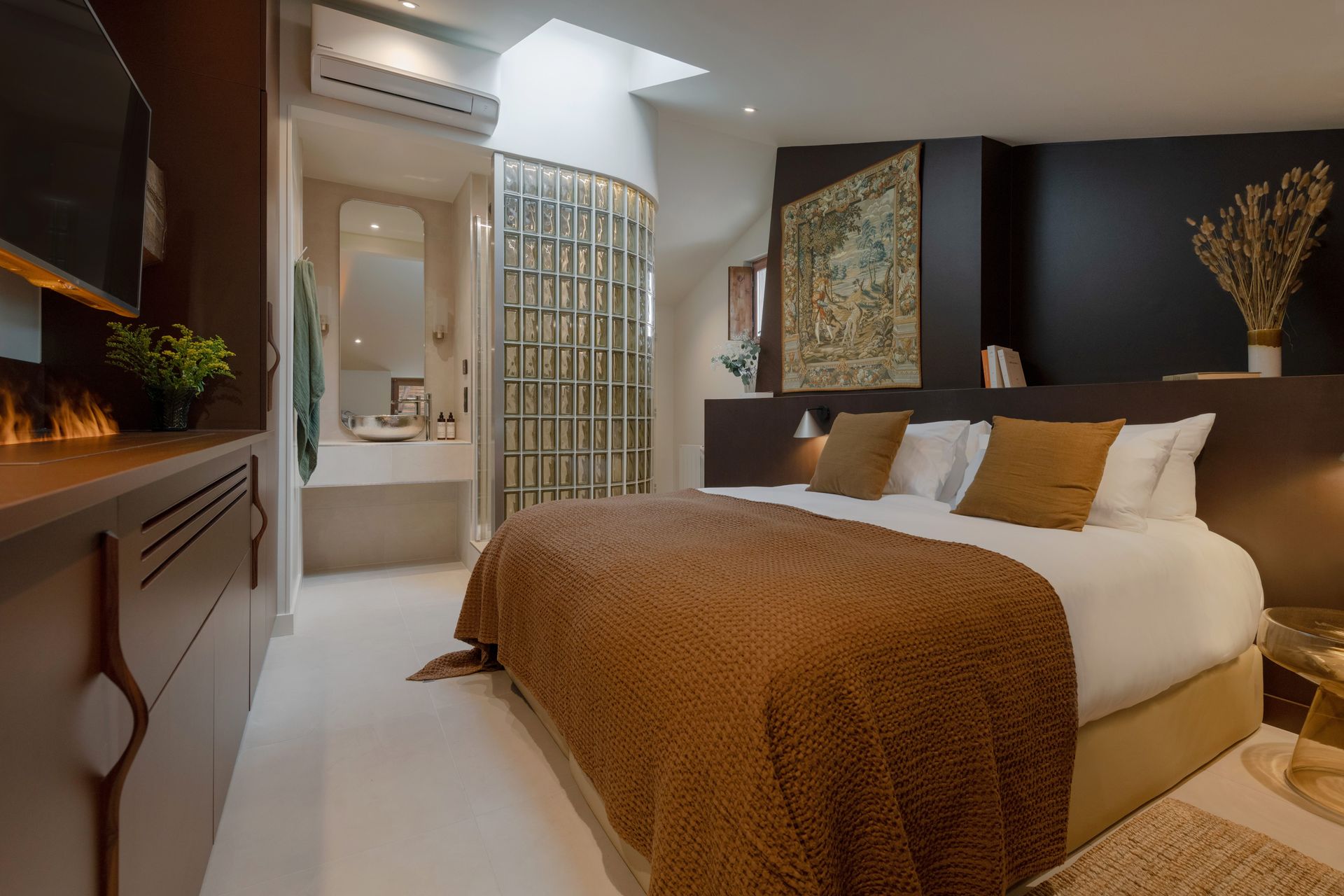 Chambre avec lit, salle de bain ouverte. Tons chauds, tapis et œuvres d'art.