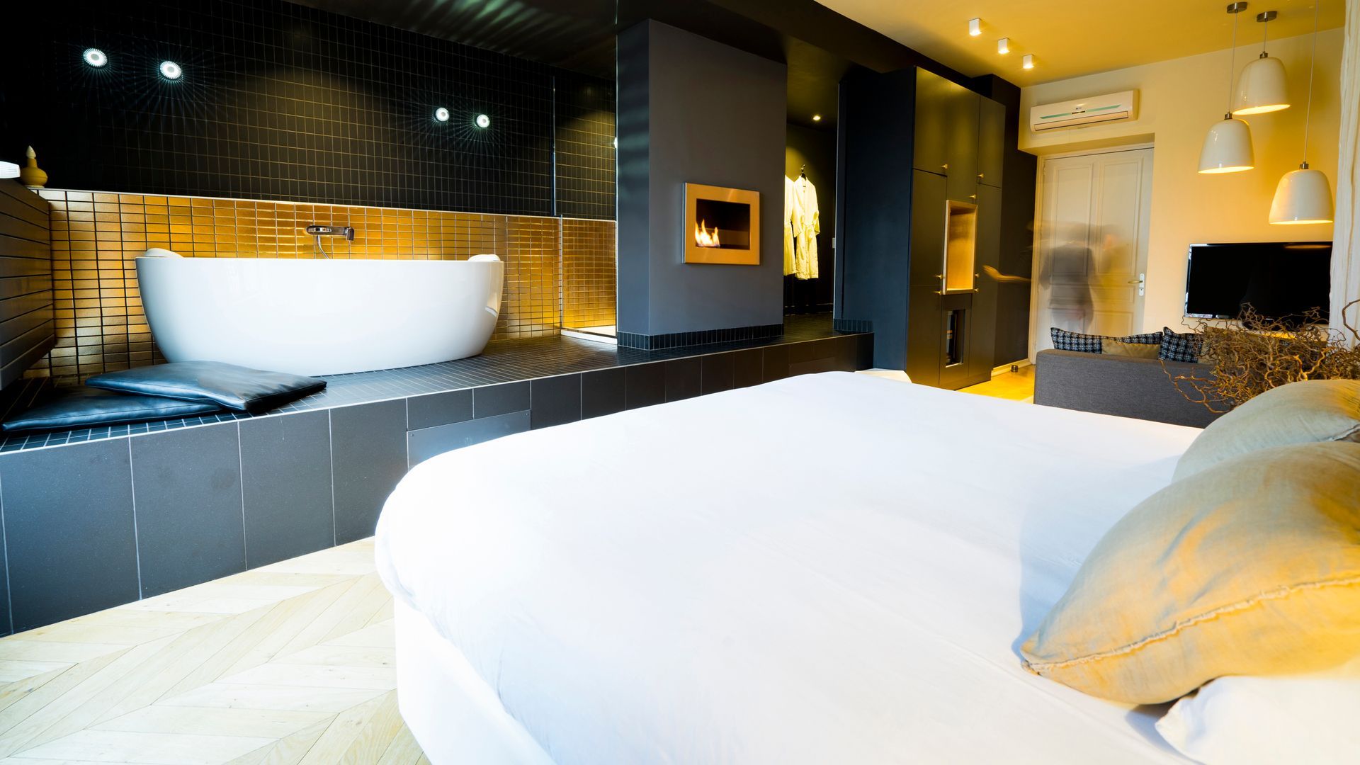 Chambre d'hôtel moderne avec lit blanc, salle de bain carrelée de noir et baignoire îlot éclairée par une douce lumière d'ambiance.