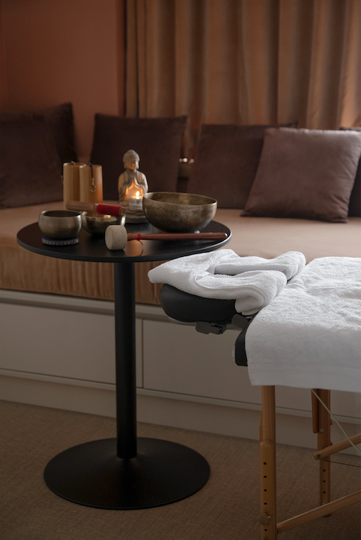 Table de massage avec serviette pliée, et table avec bols chantants et bougies, dans un décor de spa.