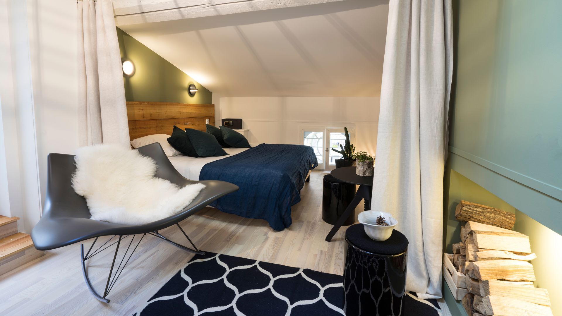 Chambre mansardée confortable avec literie bleu marine, rideaux blancs et fauteuil sur un tapis foncé.