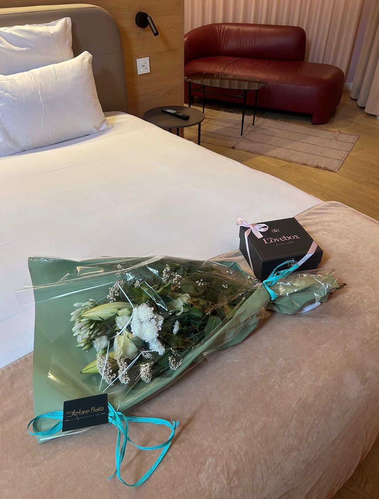 Un bouquet de fleurs et une boîte cadeau sur un lit dans une chambre d'hôtel, avec un canapé rouge en arrière-plan.