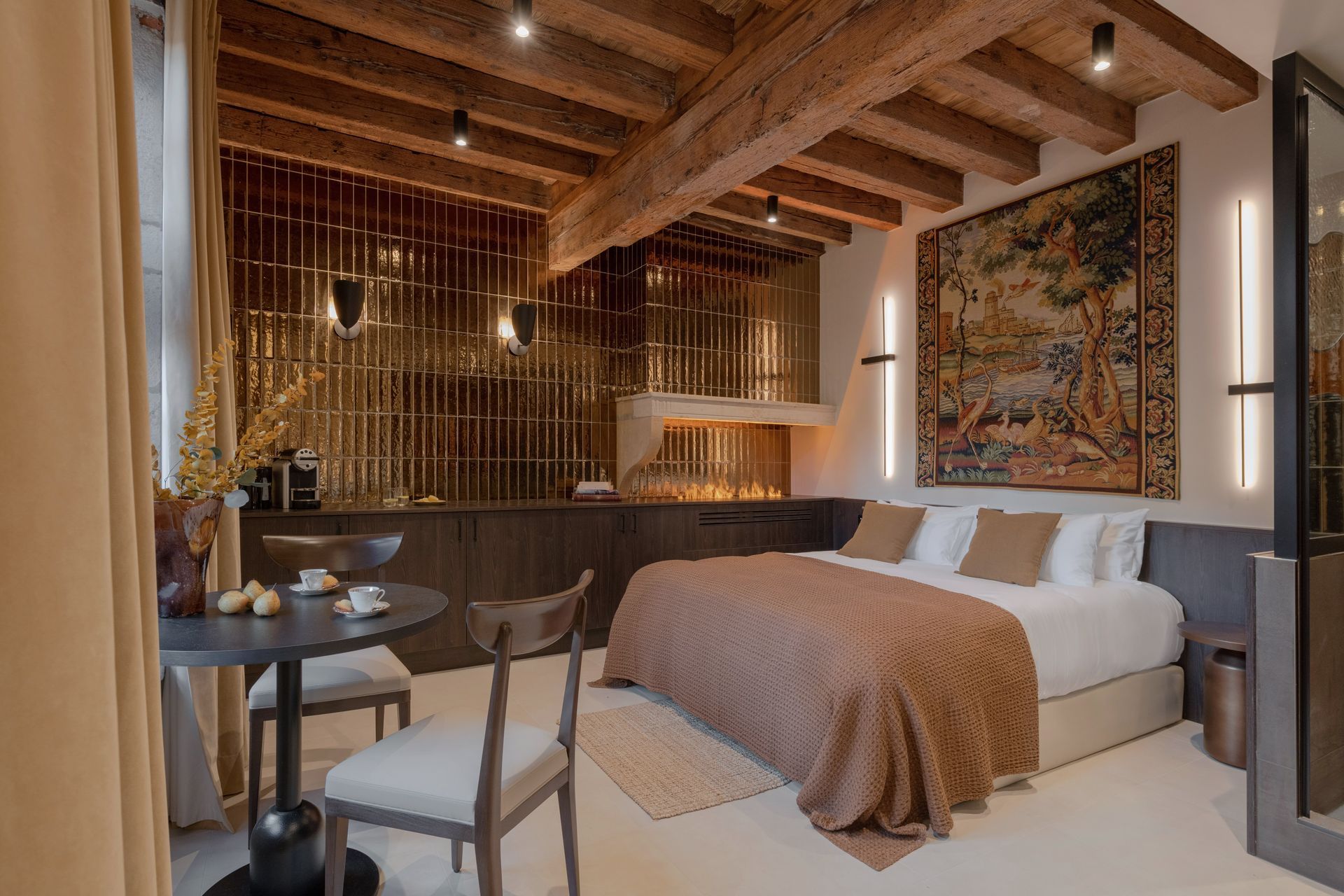 Chambre avec poutres apparentes, tapisserie, lit, table ronde et touches dorées.