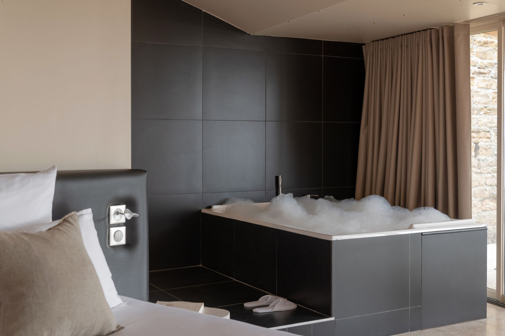 Modern bedroom with a black freestanding bathtub filled with foam beside a bed and floor-to-ceiling curtains