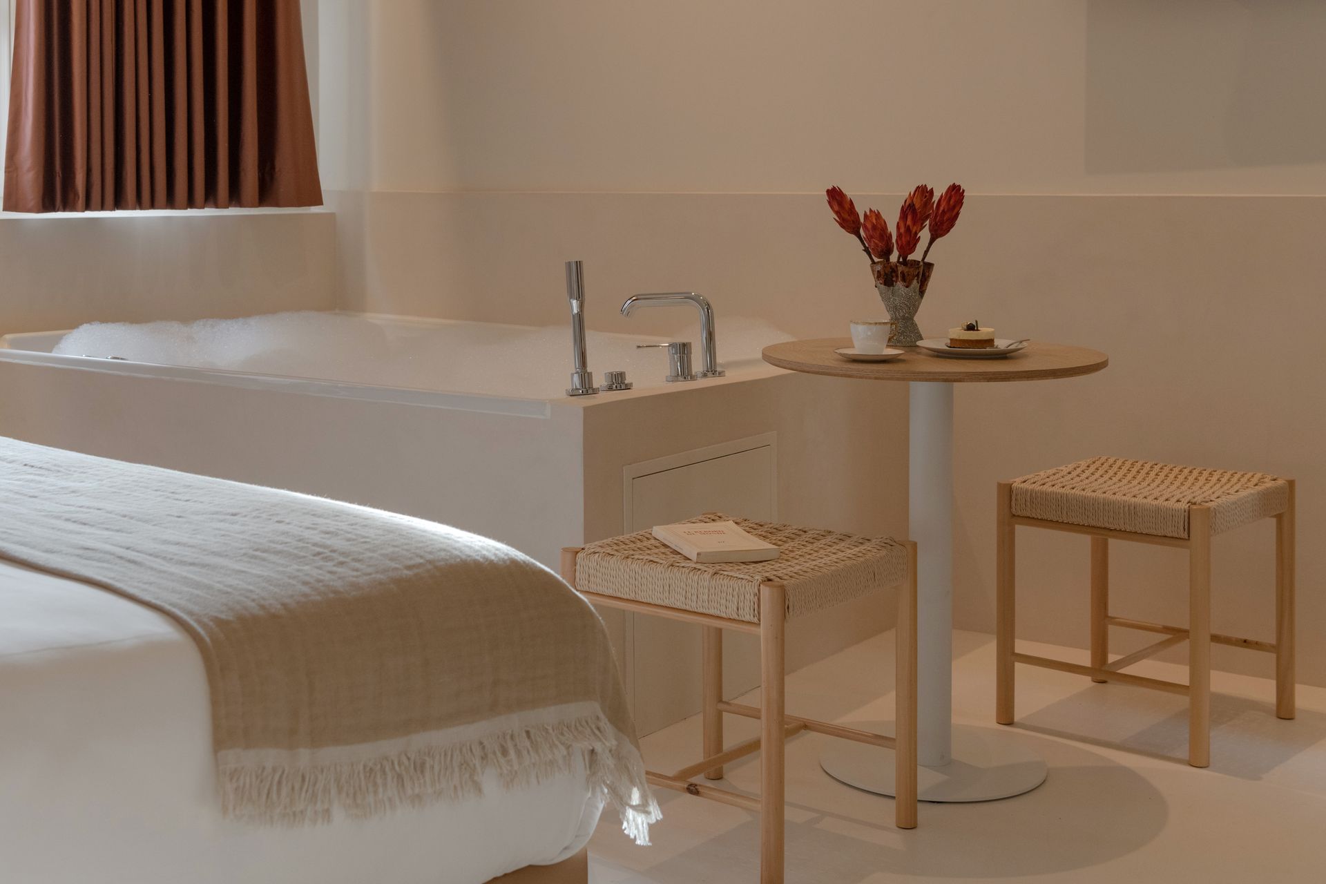 Minimal beige bedroom with a bed, wooden stools, and a small round table by a JACUZZI.