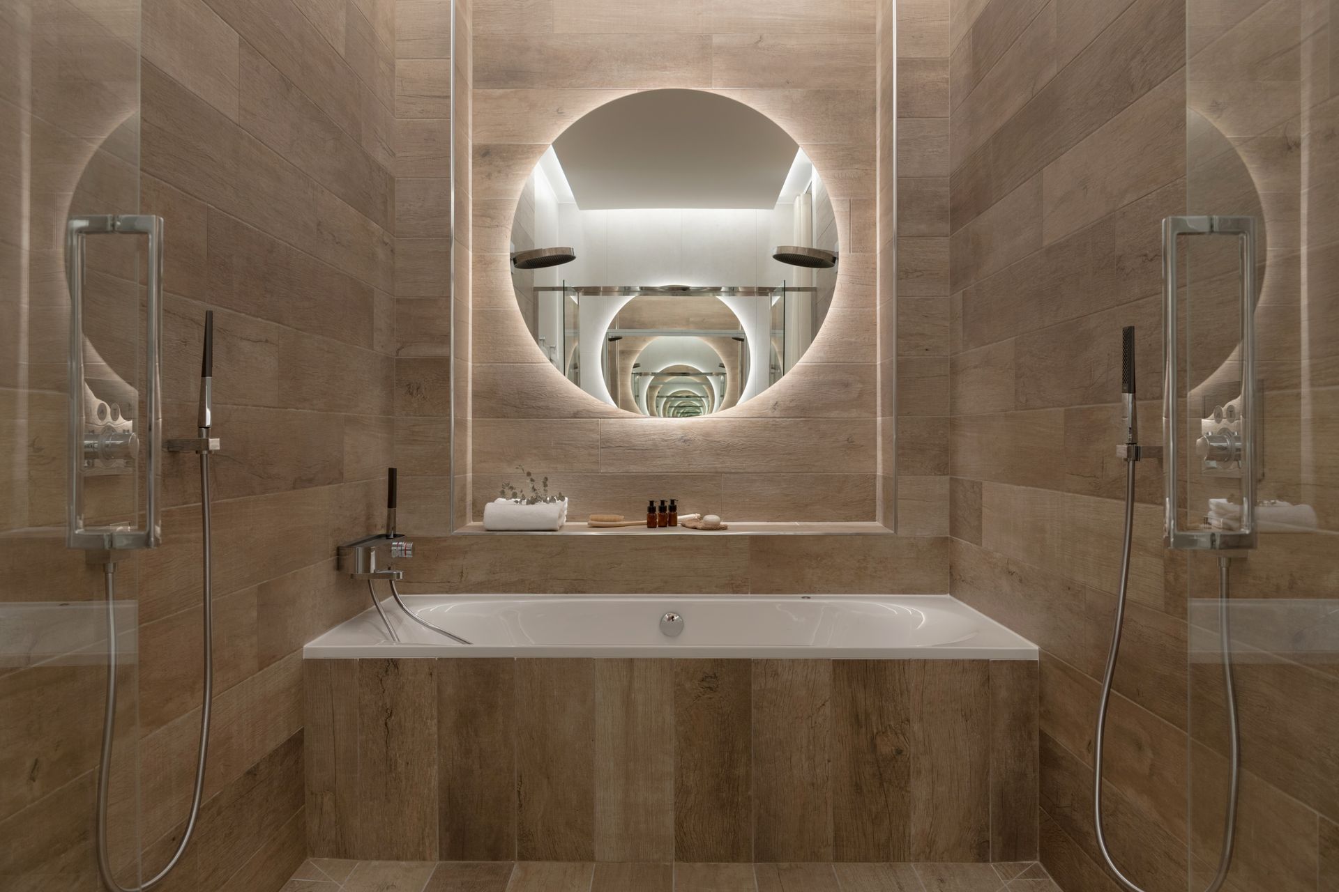 Salle de bains moderne avec baignoire, miroir rond et deux douches ; carrelage beige.