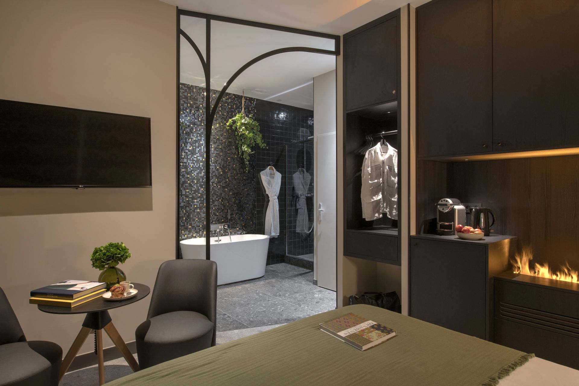 Chambre d'hôtel avec salle de bain ouverte, murs gris, lit, chaises et cheminée.