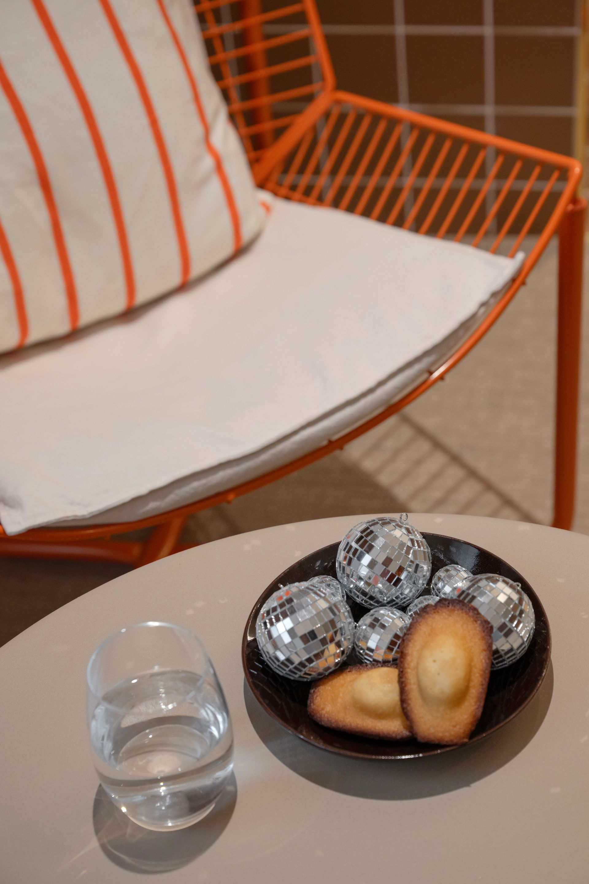 Une chaise orange à coussin rayé est placée à côté d'une table où se trouvent un verre et un bol de chocolats et de biscuits emballés.