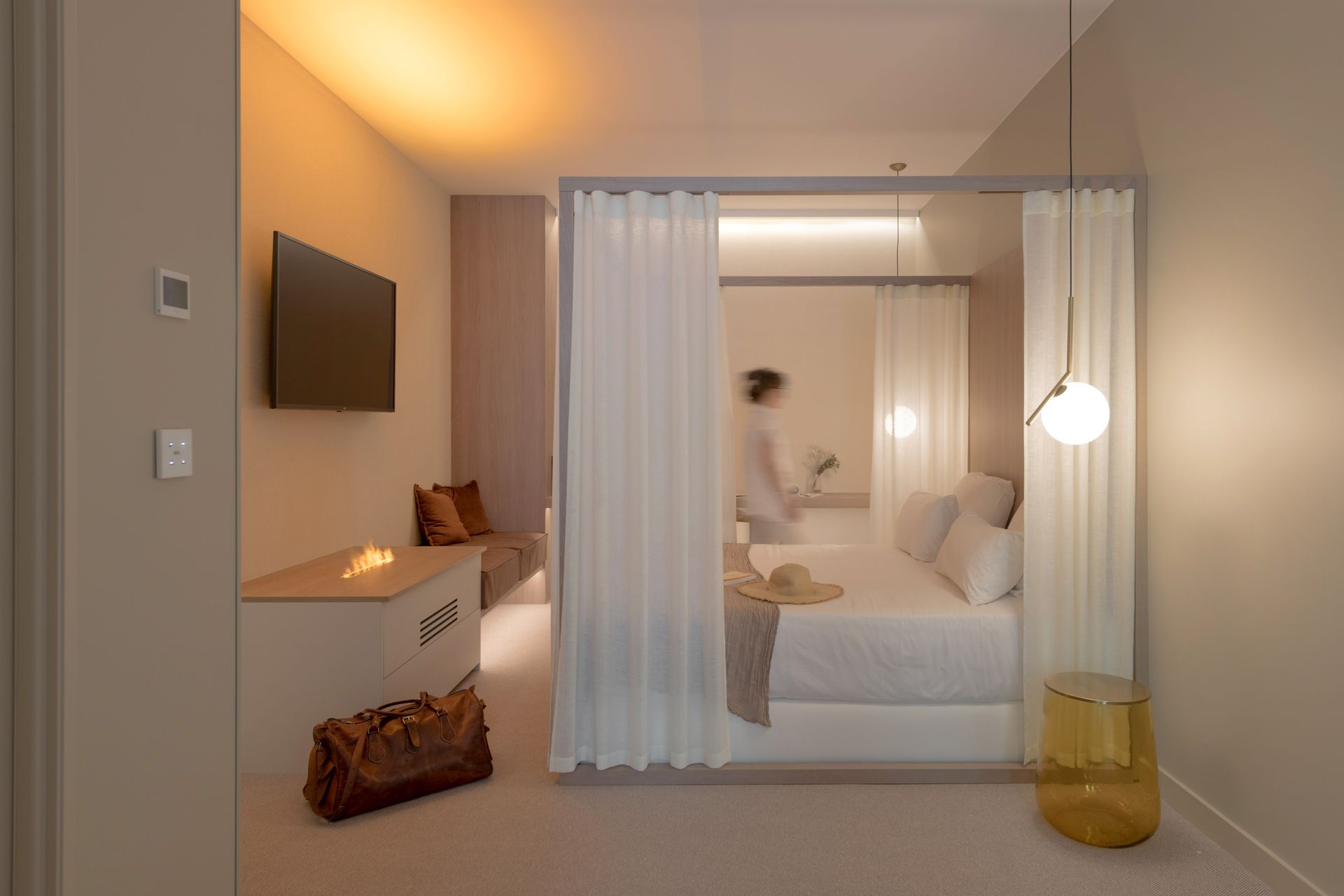 Chambre moderne avec un lit, une télévision murale, un éclairage chaleureux et des voilages séparant un coin salon.