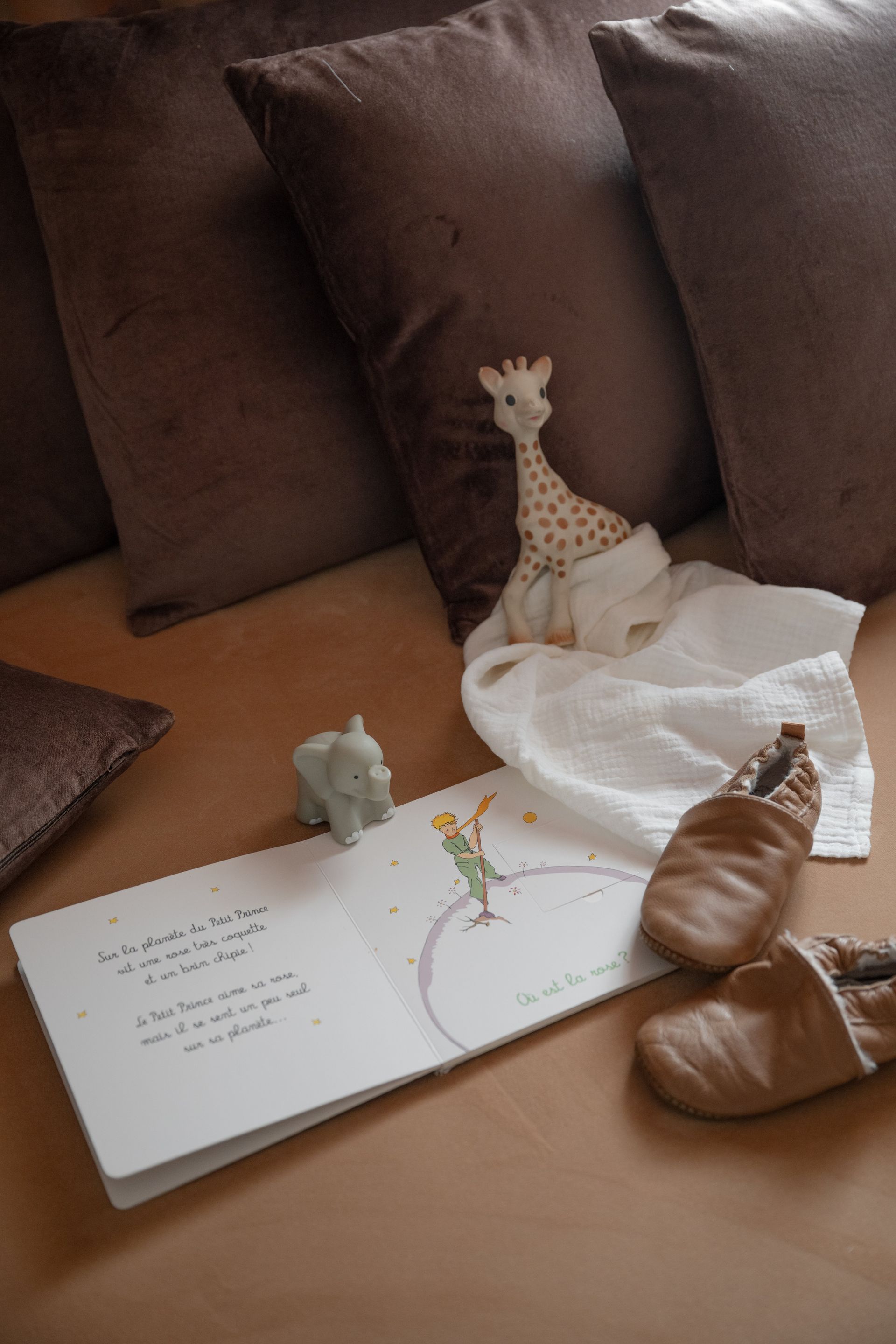 Un livre ouvert sur un canapé beige, avec à proximité une girafe, un éléphant, un lapin et des chaussures marron en peluche.