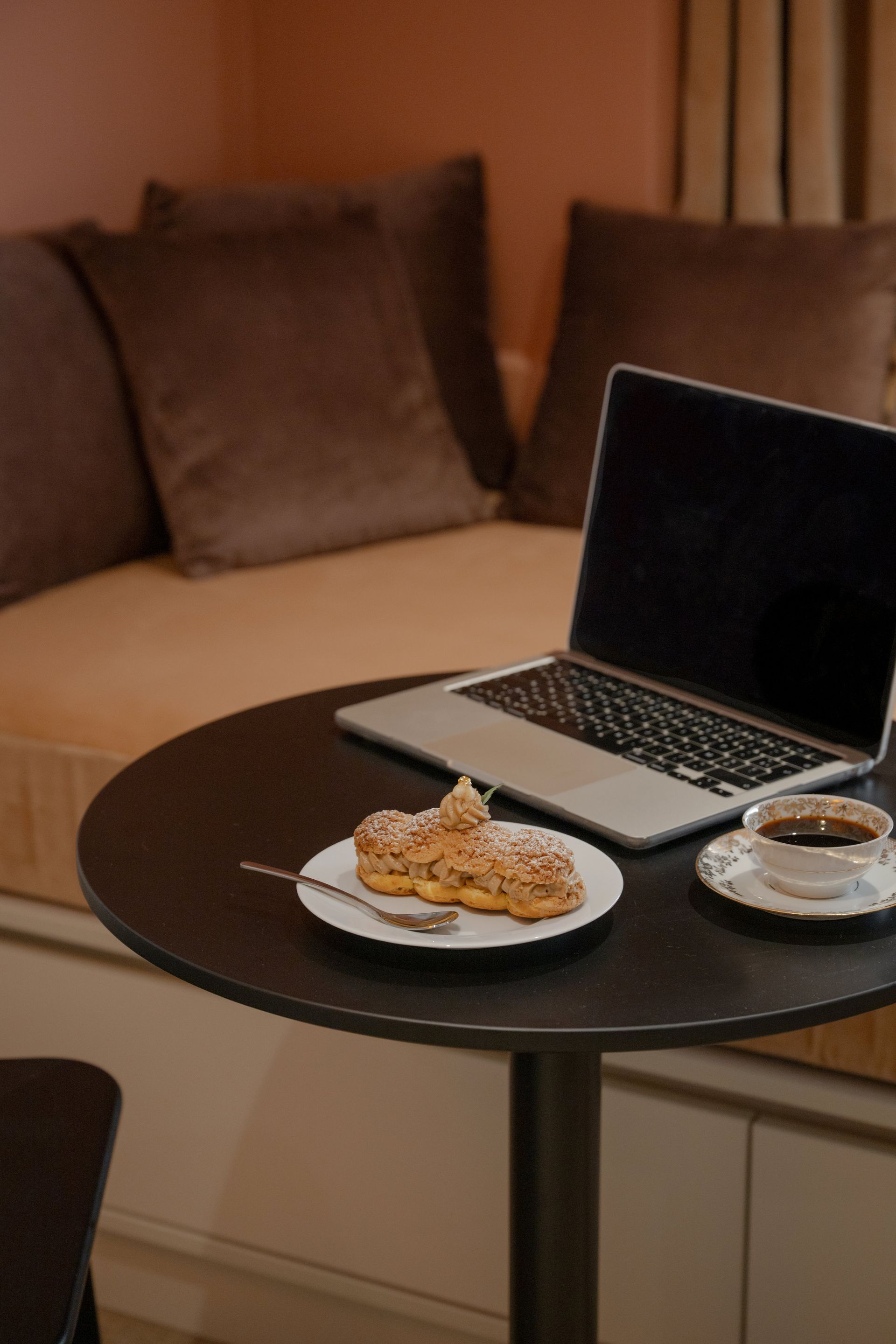 Un ordinateur portable sur une table ronde, avec une assiette de viennoiseries et une tasse dans un salon confortable.