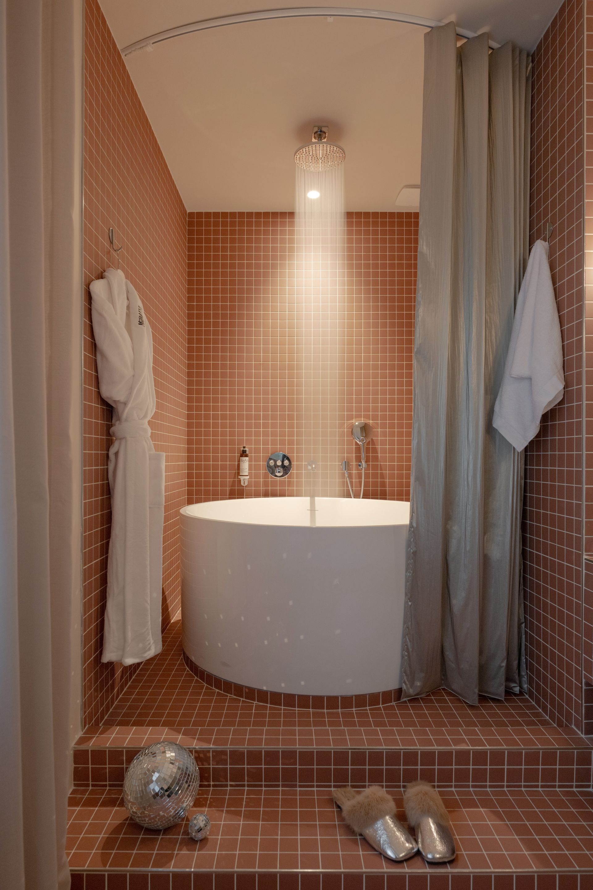 Petite salle de bains carrelée avec baignoire blanche, rideau de douche, applique murale et chaussons au sol.