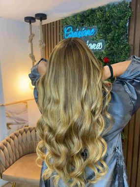 Mujer de cabello largo y ondulado en un salón de belleza, frente a un letrero de neón y una pared con plantas verdes.