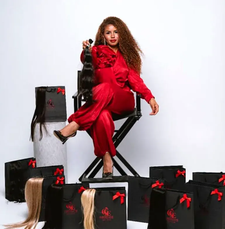 Mujer con un conjunto de satén rojo, sentada entre cajas de regalo negras con lazos rojos, sosteniendo una pieza de cabello postizo