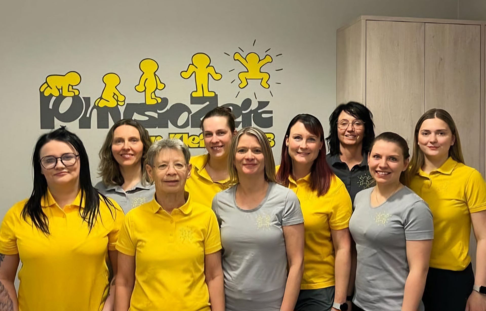 Gruppe lächelnder Frauen in gelben Hemden, die vor einem Logo an einer Wand stehen.